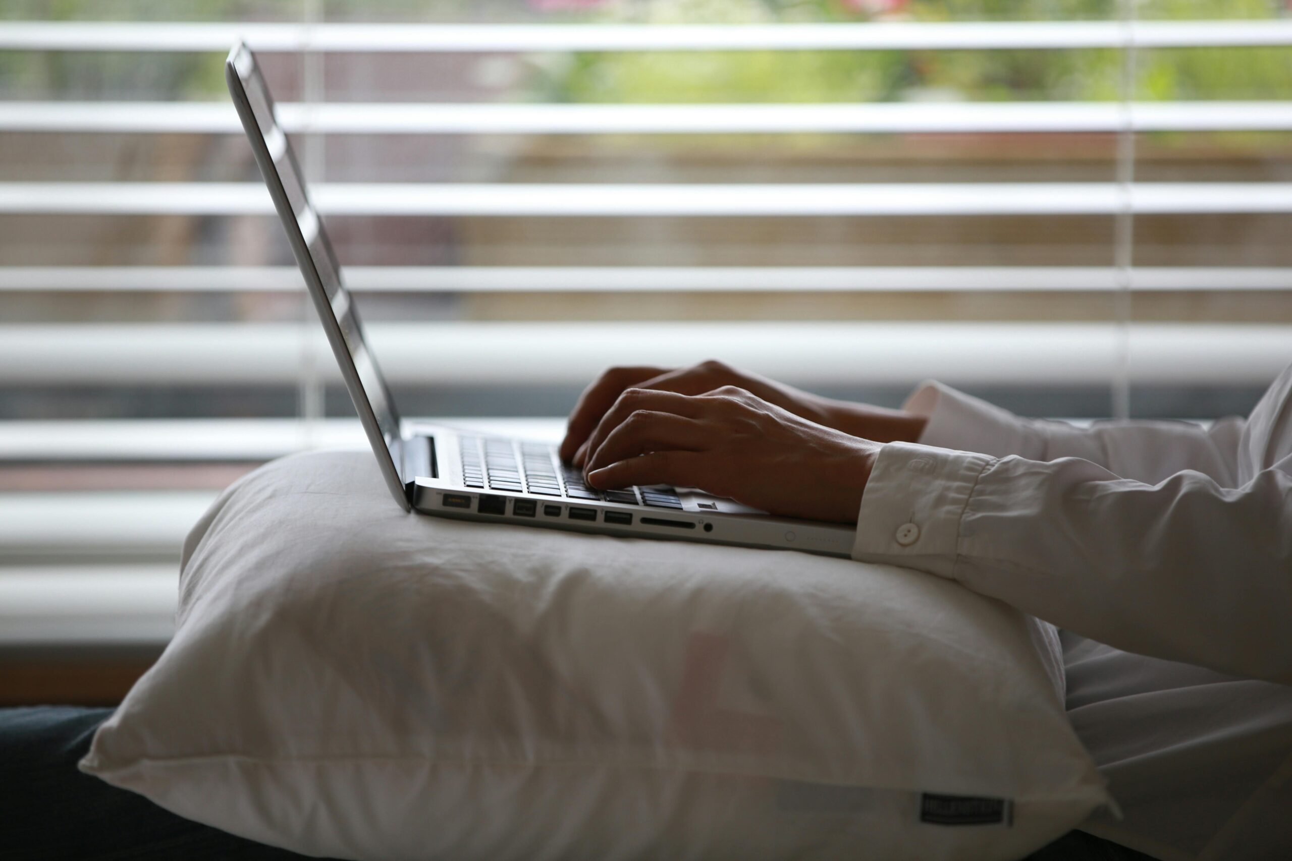 Close-up of hands typing on a laptop while seated on a pillow indoors.