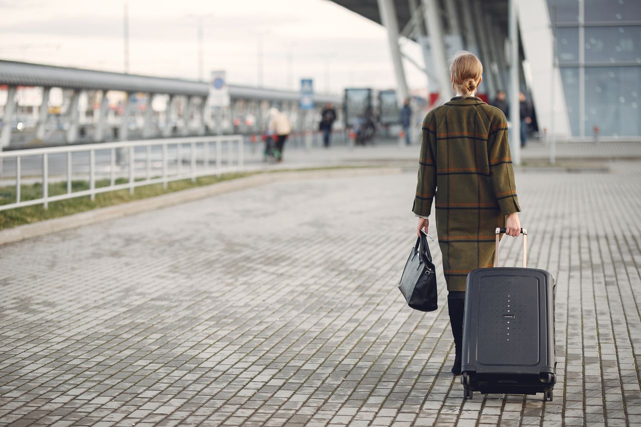 Back view full length of female executive in trendy outfit with luggage walking along sidewalk into airport building before departure